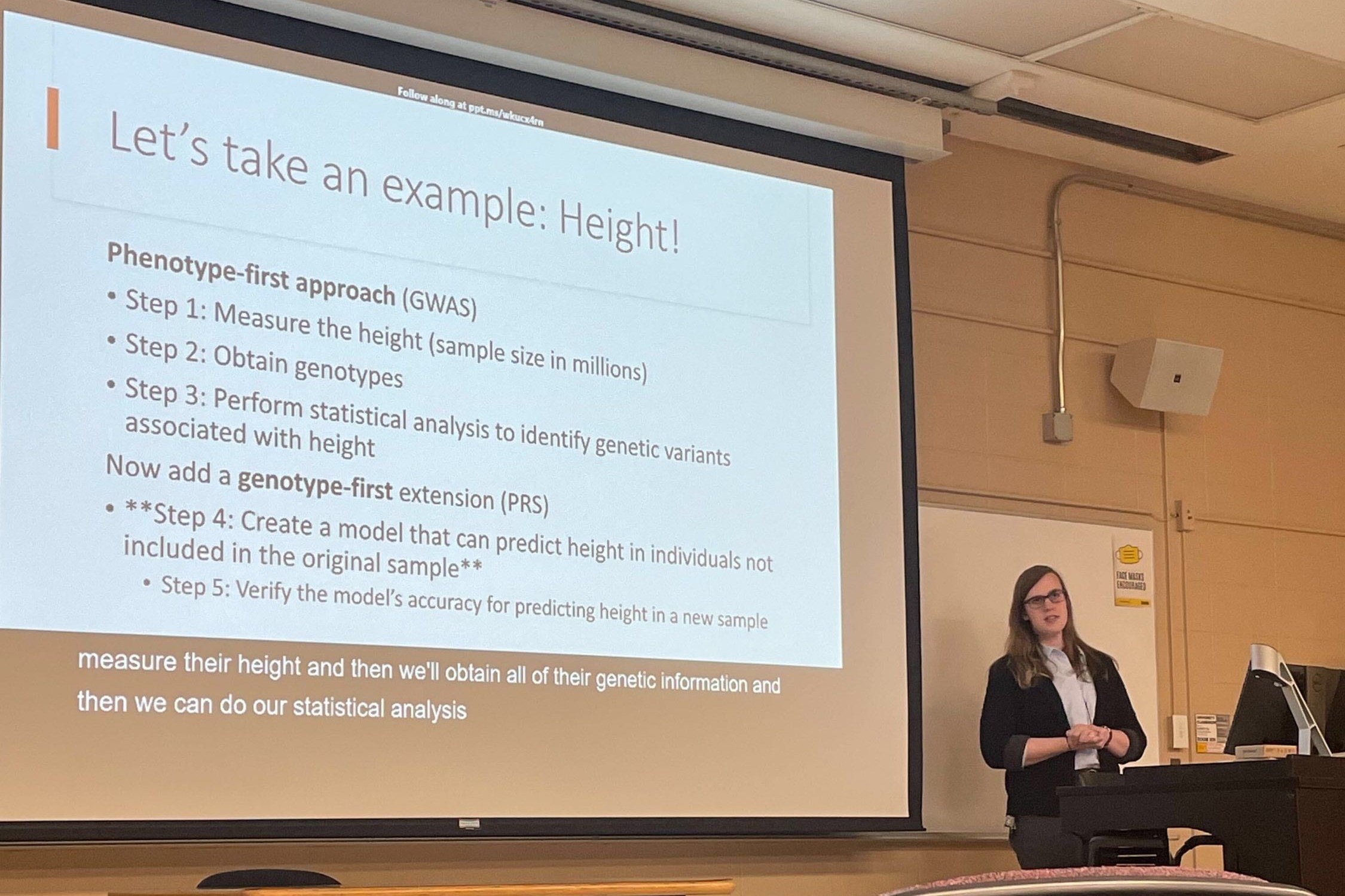 Photograph of the website's owner, Valerie, presenting a talk at the University of Iowa. The image shows Valerie alongside a Powerpoint presentation, with the current slide using Height to explain polygenic traits.